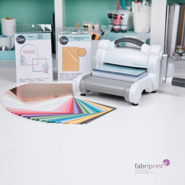 Guilhotina Sizzix Big Shot com acessórios e papéis coloridos sobre uma mesa de trabalho.